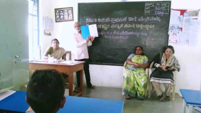 Retired teacher addressing students at a government school in Muddenahalli during Dr. H. Narasimhaiah’s birthday celebration with a portrait of Dr. Narasimhaiah and Gandhi in the background. - Local News